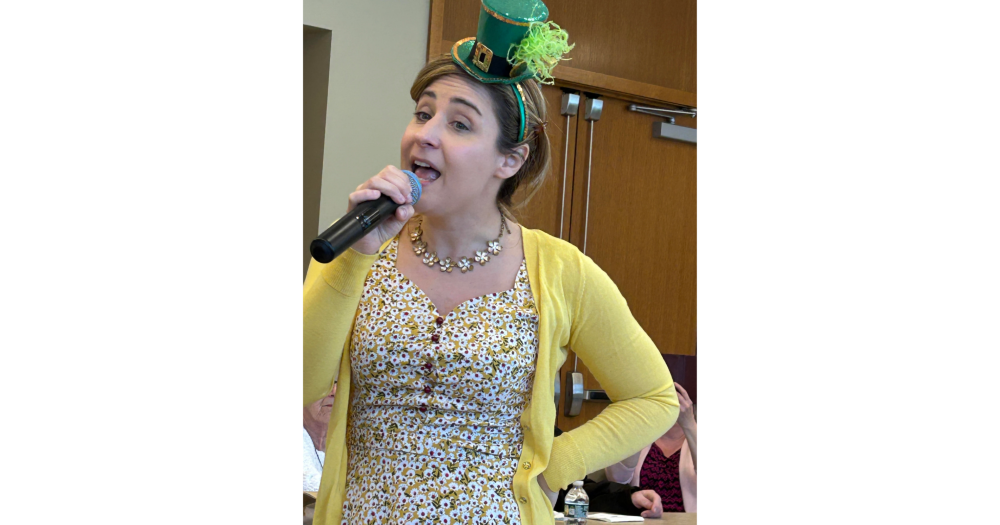 A woman with a microphone entertaining memory cafe guests on 3-7-2025 while wearing a leprechaun hat.